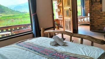 A room with a bed, a wooden headboard, a window with a view of green hills, and a brick wall.