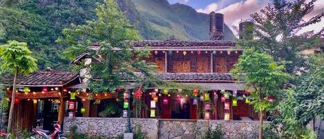 A traditional wooden building with a stone foundation, surrounded by lush greenery and mountains in the background.