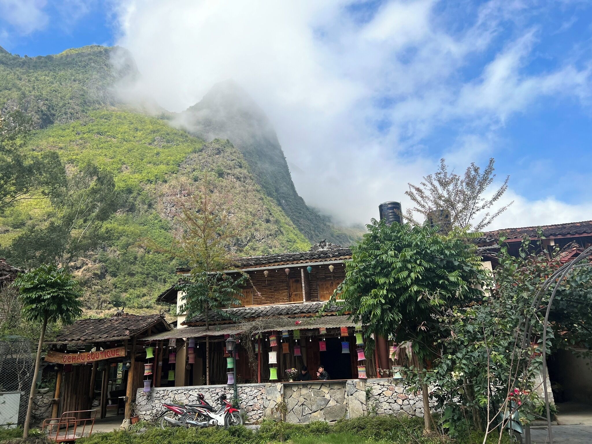 Edificio in legno tradizionale con parete in pietra, una motocicletta parcheggiata davanti e una montagna con nuvole sullo sfondo.