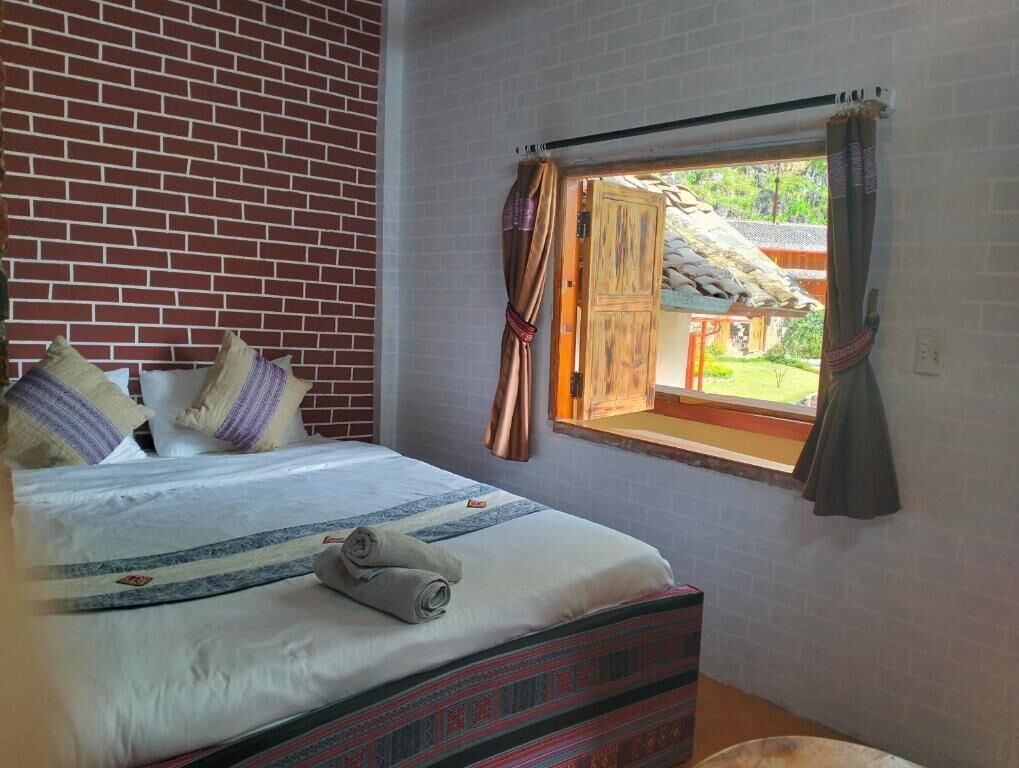 A bedroom with a brick wall, a bed with patterned bedding, a window with curtains, and a view of an outdoor area.