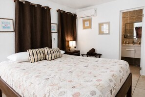 Habitación de hotel con cama, mesita de noche con lámpara, una silla y baño a la vista a través de una puerta abierta.