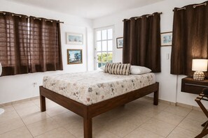 A bedroom with a bed, curtains, a bedside table with a lamp, and two framed pictures on the wall.