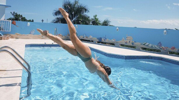 Une personne plonge dans une piscine à l’eau claire et bleue, dont le bord est décoratif, avec un bâtiment visible en arrière-plan.