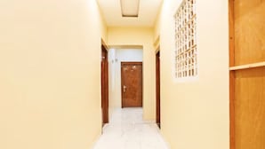 A narrow hallway with a wooden door at the end, a window with a grid pattern, and a white tiled floor. - Vrrinda By GRB (Varanasi)
