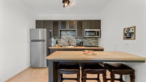 Eine moderne Küche mit Edelstahl-Kühlschrank, einer Mikrowelle und einer gefliesten Spritzschutzwand. Eine Holzinsel mit Sitzgelegenheiten und einer Schüssel mit Obst ist ebenfalls vorhanden.