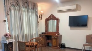 Une chambre à coucher comprenant un lit, une commode en bois, un bureau, une chaise, un téléviseur et un grand miroir.