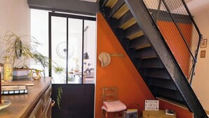 A room with a wooden dresser, a pink chair, a small table with a lamp, a staircase with a black railing, and a door leading to another room. - Cosy, country-style 3* gîte in the heart of Picardy's heritage, sleeps 5 (Mesbrecourt-Richecourt)