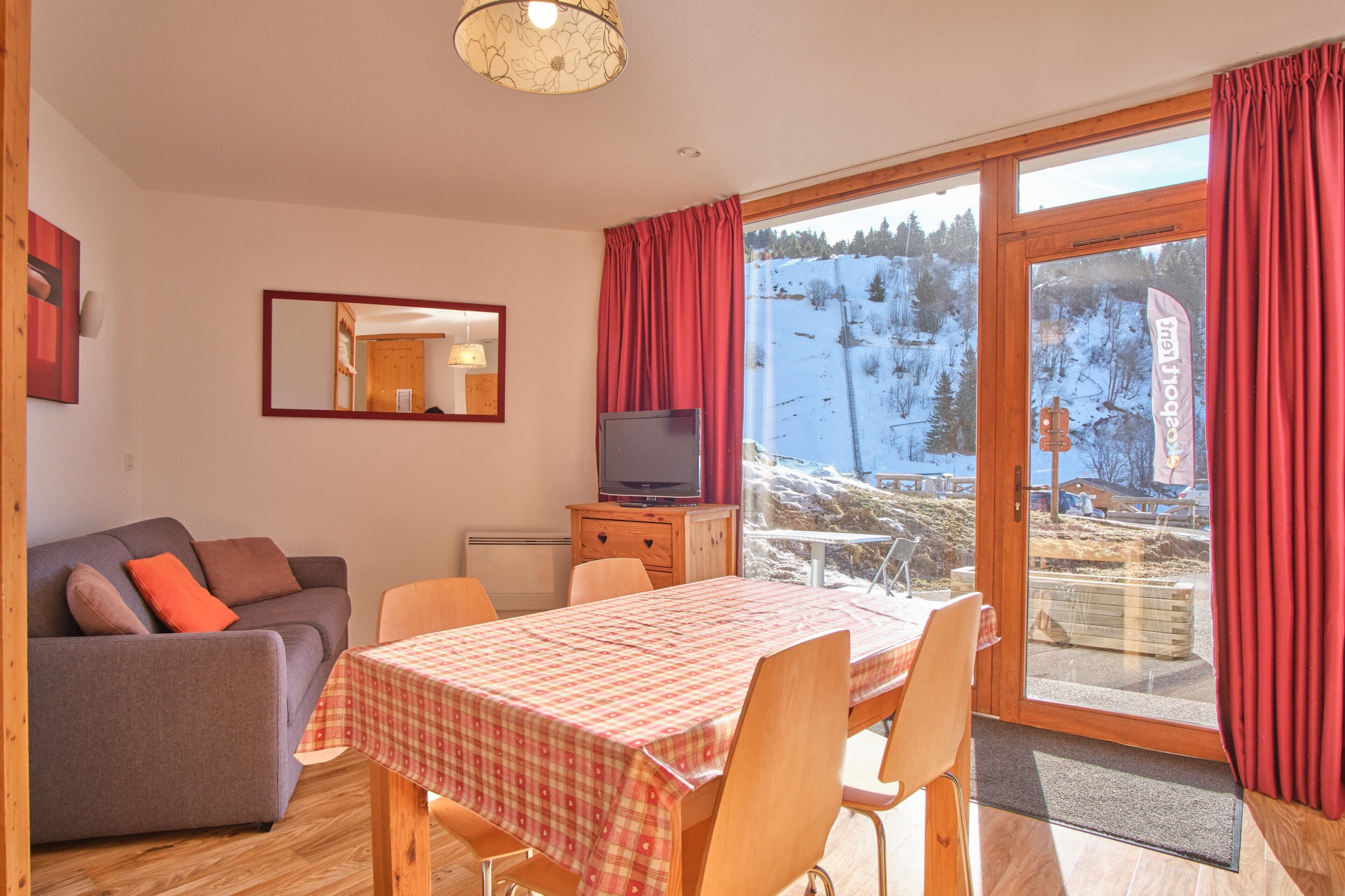 Une pièce comprenant un canapé, une table à manger avec des chaises, un téléviseur et une grande fenêtre avec des rideaux.