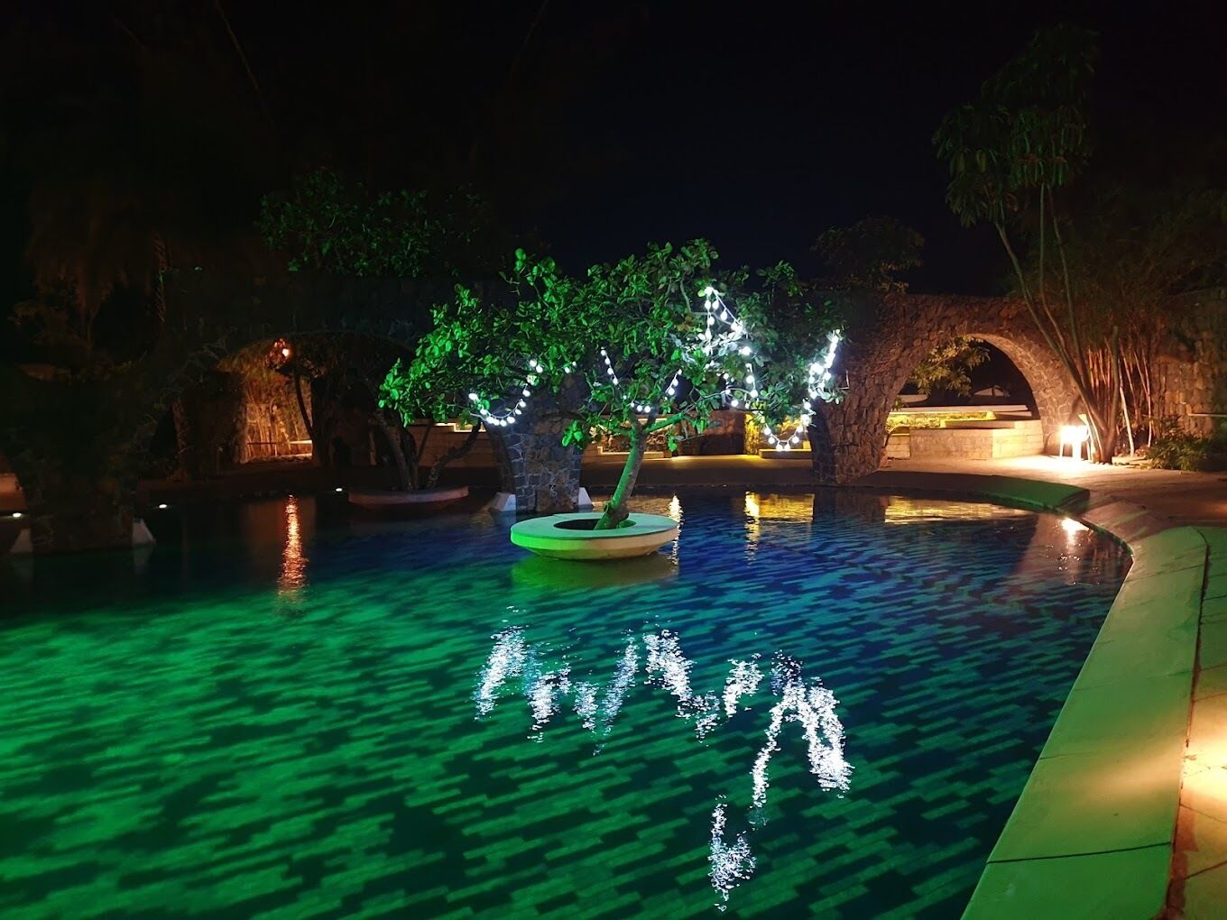 A nighttime pool area with a tree illuminated by string lights, surrounded by stone structures and a stone bridge.