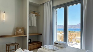 Habitación de hotel con dos camas, una estantería de madera, un taburete pequeño, una maleta y vistas al mar.