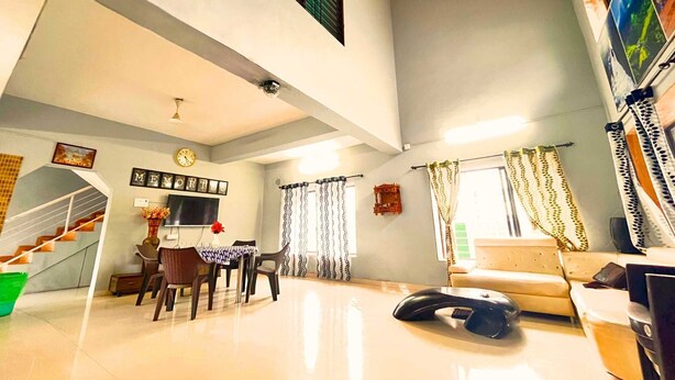 A spacious living area with a staircase, a dining table with chairs, a sofa, and a television mounted on the wall.
