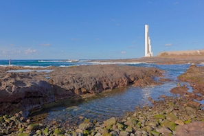 A rocky shoreline with clear water, a tall white structure, and a distant building.
