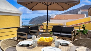 Una terrazza con un tavolo apparecchiato per due, un cesto di vimini con limoni e vista sul mare e sulle montagne.