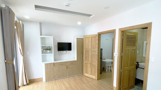 A bedroom with a bed, wooden wardrobe, a TV mounted on the wall, and a bathroom visible through an open door. - Jolilian Hotel & Apartment (Ho Chi Minh City)