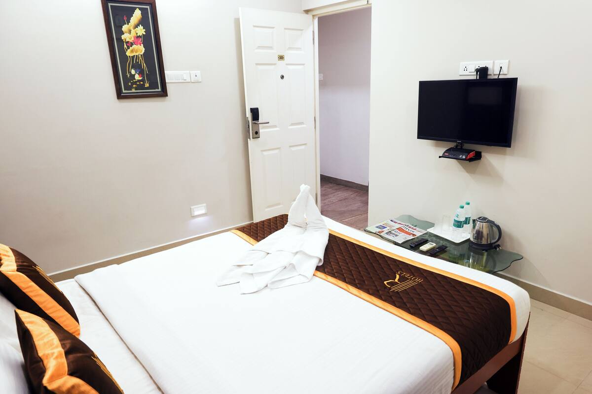 A hotel room with a bed, a TV mounted on the wall, a bedside table with a kettle, and a door leading to another room.