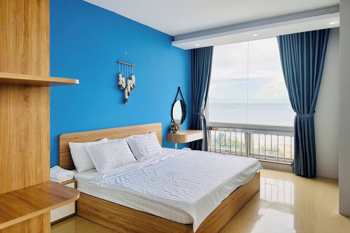 A modern bedroom with a large bed, a wooden nightstand, a bedside table, a wall-mounted mirror, and a view of the sea through a balcony.