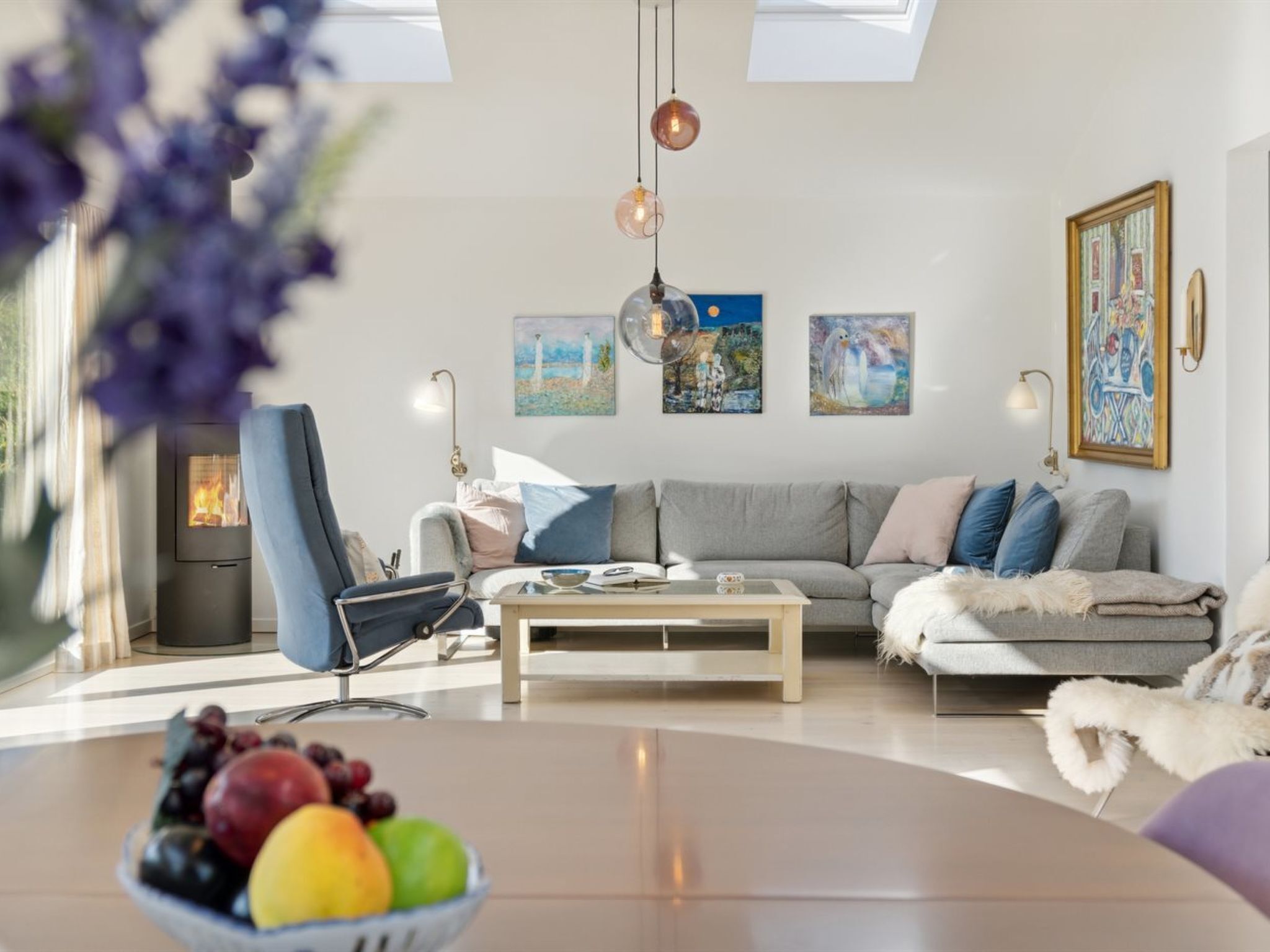 Una sala de estar moderna con sofá, sillón y mesa de centro. Hay una chimenea, obras de arte en la pared y un frutero sobre la mesa.
