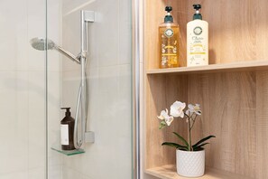Um chuveiro com chuveiro manual e torneira fixada na parede, uma garrafa de sabonete líquido e uma garrafa de shampoo em uma prateleira de madeira.