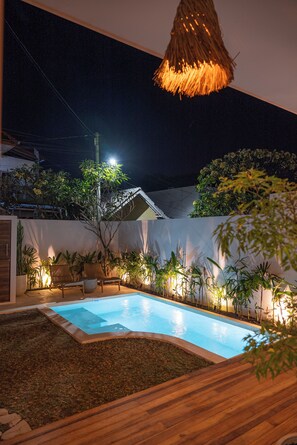 Un espace piscine aménagé pour la nuit, avec un luminaire en forme de toiture de chaume, entouré de plantes en pot et doté d’une terrasse en bois.