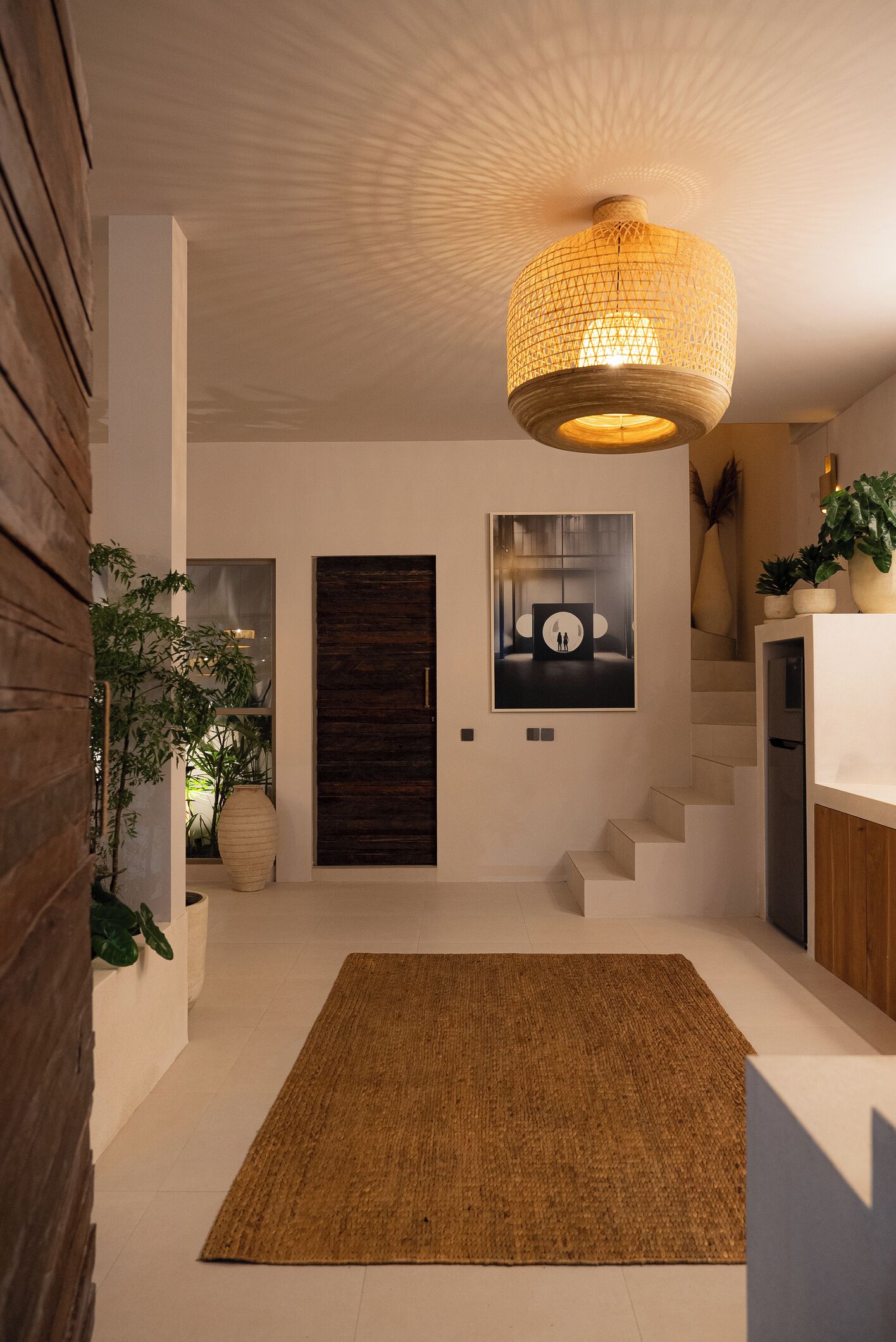 A modern interior with a large woven light fixture, a dark wooden door, a framed artwork, and a staircase.