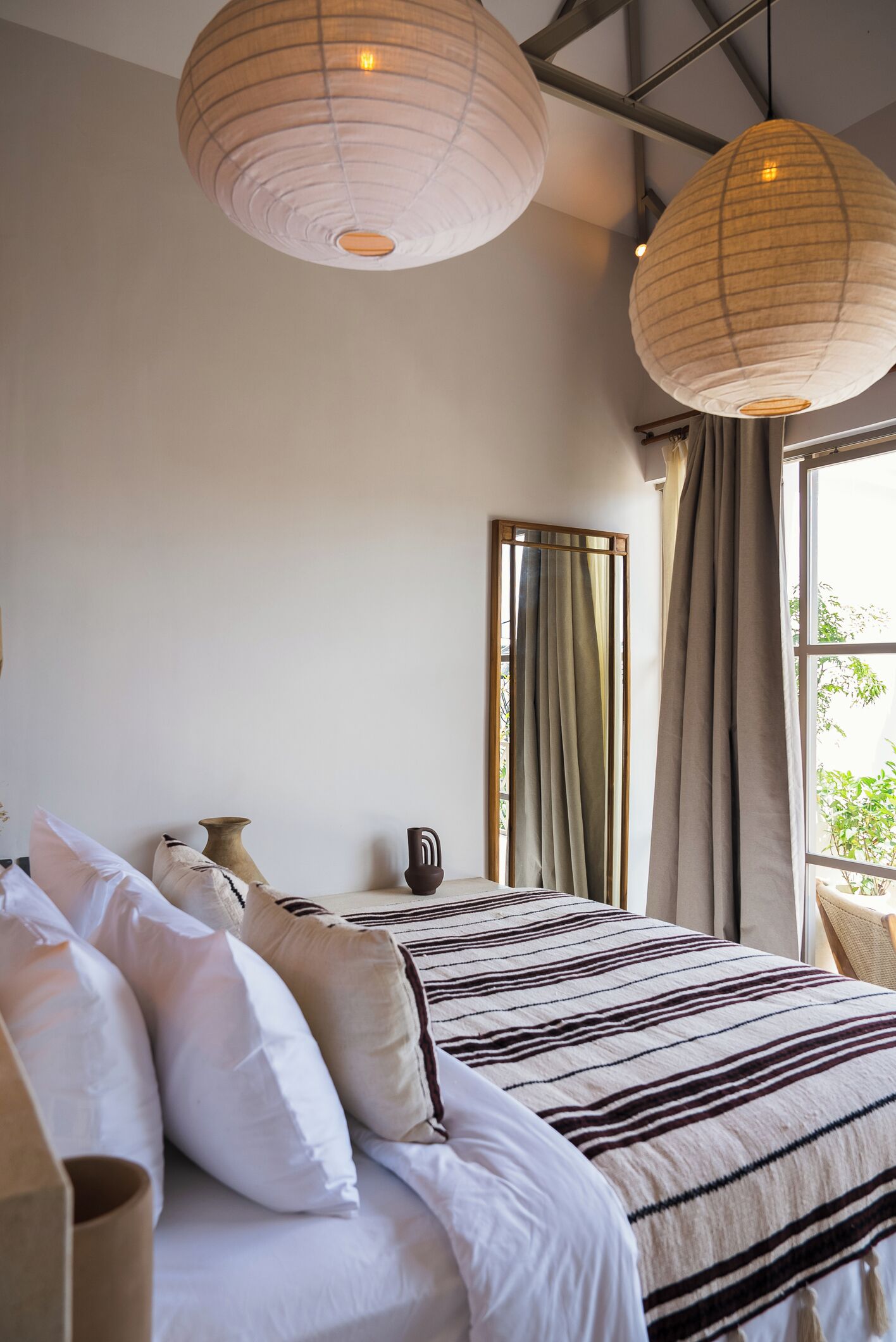 A bedroom with a large bed, multiple pillows, a bedside table, a mirror, and two hanging paper lanterns.