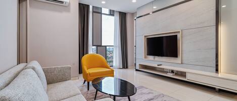 Un salon moderne avec un canapé, un fauteuil jaune, une table basse en verre, un téléviseur à écran plat et une grande fenêtre avec des rideaux.