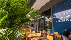 Un espacio al aire libre con mesas y sillas de madera, rodeado de exuberantes plantas verdes. Se ve un cartel grande con el texto "True to Great Coffee & Good People".