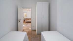 A narrow hallway with two beds, a wardrobe, and a door leading to a living area with a sofa and lamp.