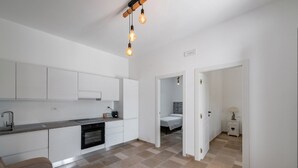 A modern kitchen with white cabinets, a countertop, and a dining area with a striped sofa. There is a bed in the adjacent room and a nightstand with a lamp.
