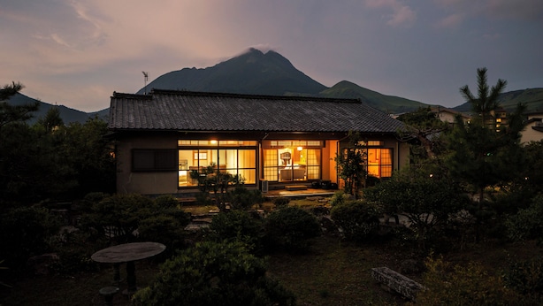 Een traditioneel Japans huis met pannendak, omringd door weelderige begroeiing en een berg op de achtergrond.