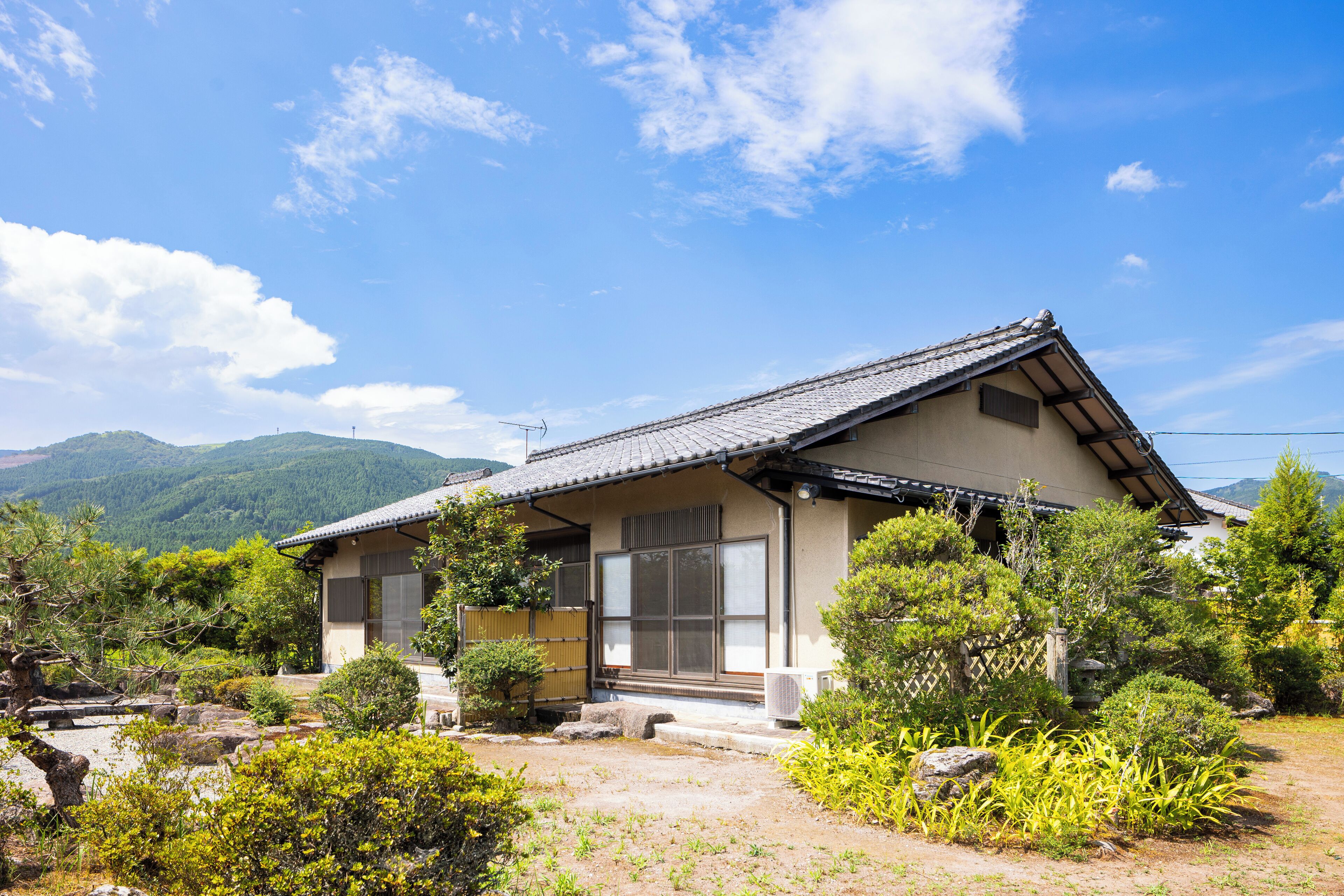 A traditional Japanese house with a tiled roof, surrounded by lush greenery and mountains in the background.