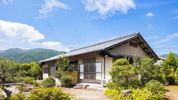A traditional Japanese house with a tiled roof, surrounded by lush greenery and mountains in the background.