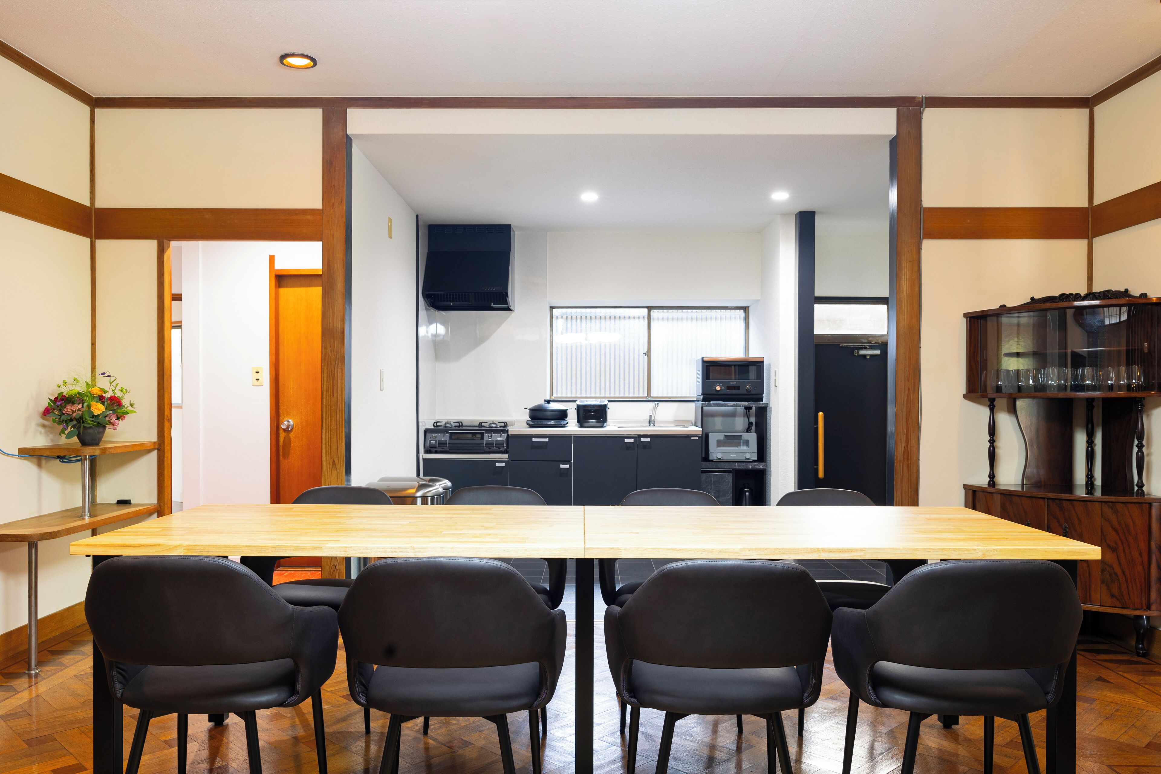 Un comedor con una mesa de madera y sillas negras, una cocina al fondo y un armario con cristalería a la derecha.