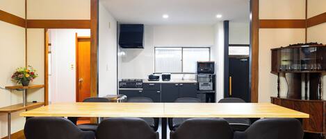 A dining area with a wooden table and black chairs, a kitchen in the background, and a cabinet with glassware on the right.