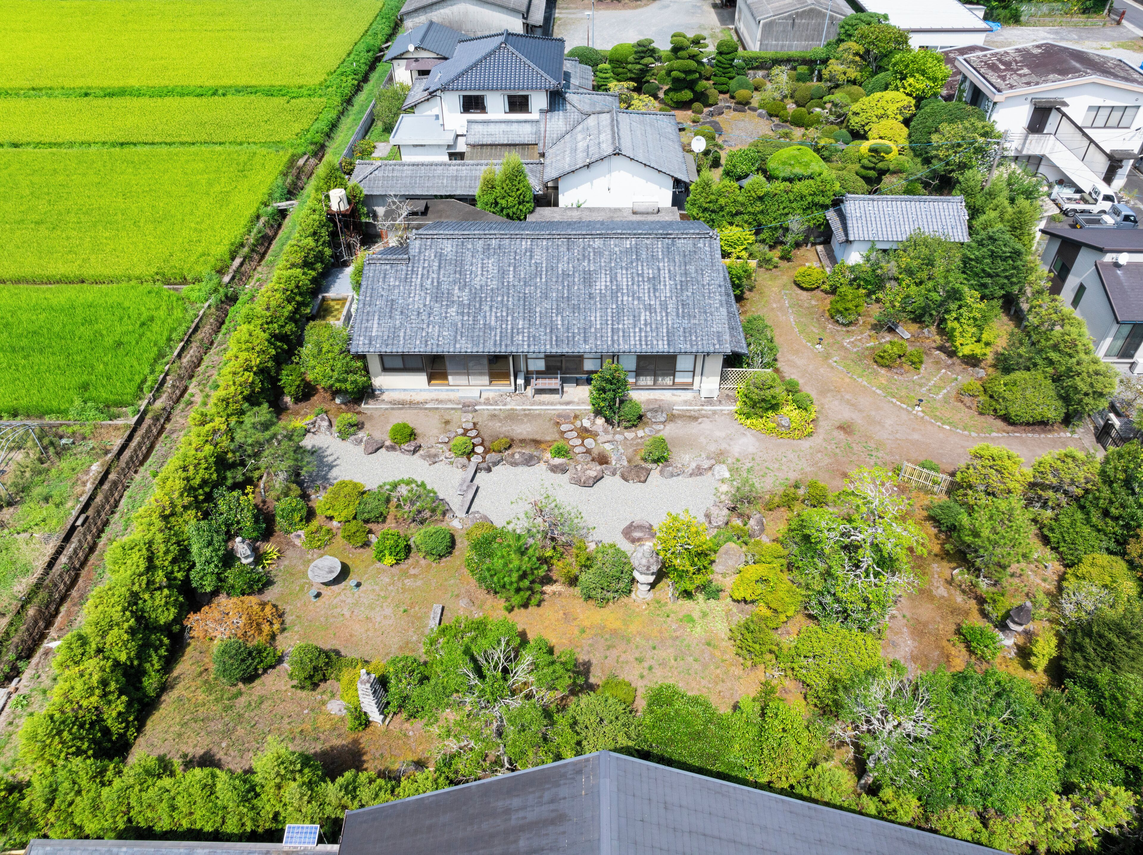 An aerial view of a residential area with traditional Japanese houses, a well-maintained garden, and adjacent agricultural fields.