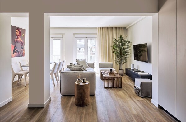 Un salon moderne avec un canapé, une table basse et un téléviseur. Il y a une grande fenêtre avec des rideaux, une plante et un tableau au mur.