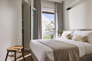 Une chambre avec un grand lit, un tabouret en bois et une vue sur un balcon donnant sur des immeubles.