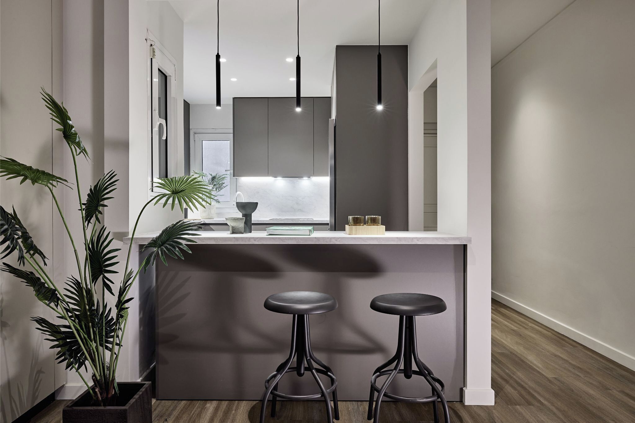 A modern kitchen with a central island, two bar stools, pendant lighting, and a potted plant.