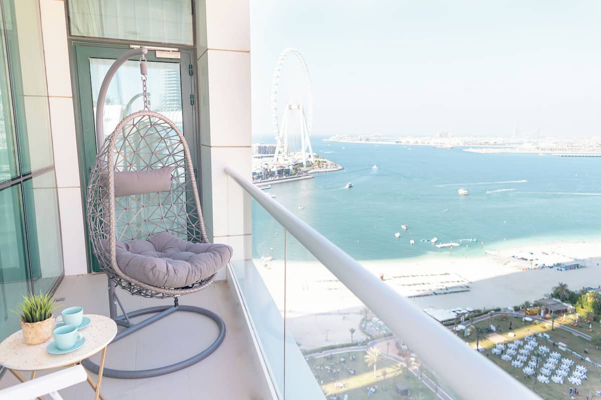 Un balcon avec un fauteuil suspendu, une petite table ornée d’une plante et de quelques tasses, et une vue sur une zone côtière avec un pont et des bateaux.
