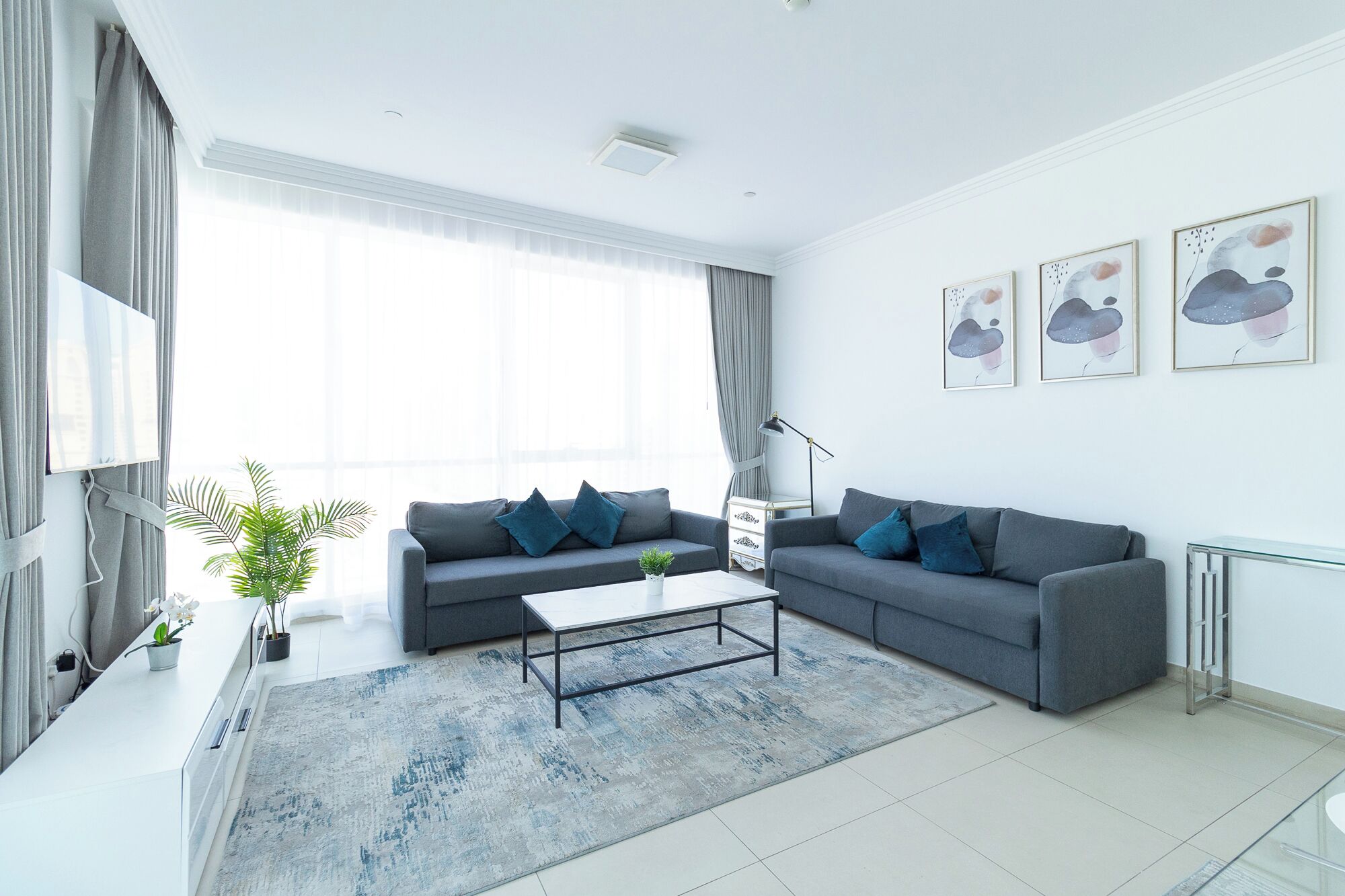 A modern living room with a grey sofa, a coffee table, and a large window with curtains. There are three framed artworks on the wall and a potted plant on the side table.
