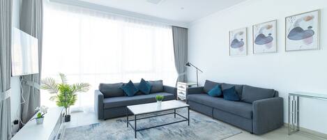 A modern living room with a grey sofa, a coffee table, and a large window with curtains. There are three framed artworks on the wall and a potted plant on the side table.