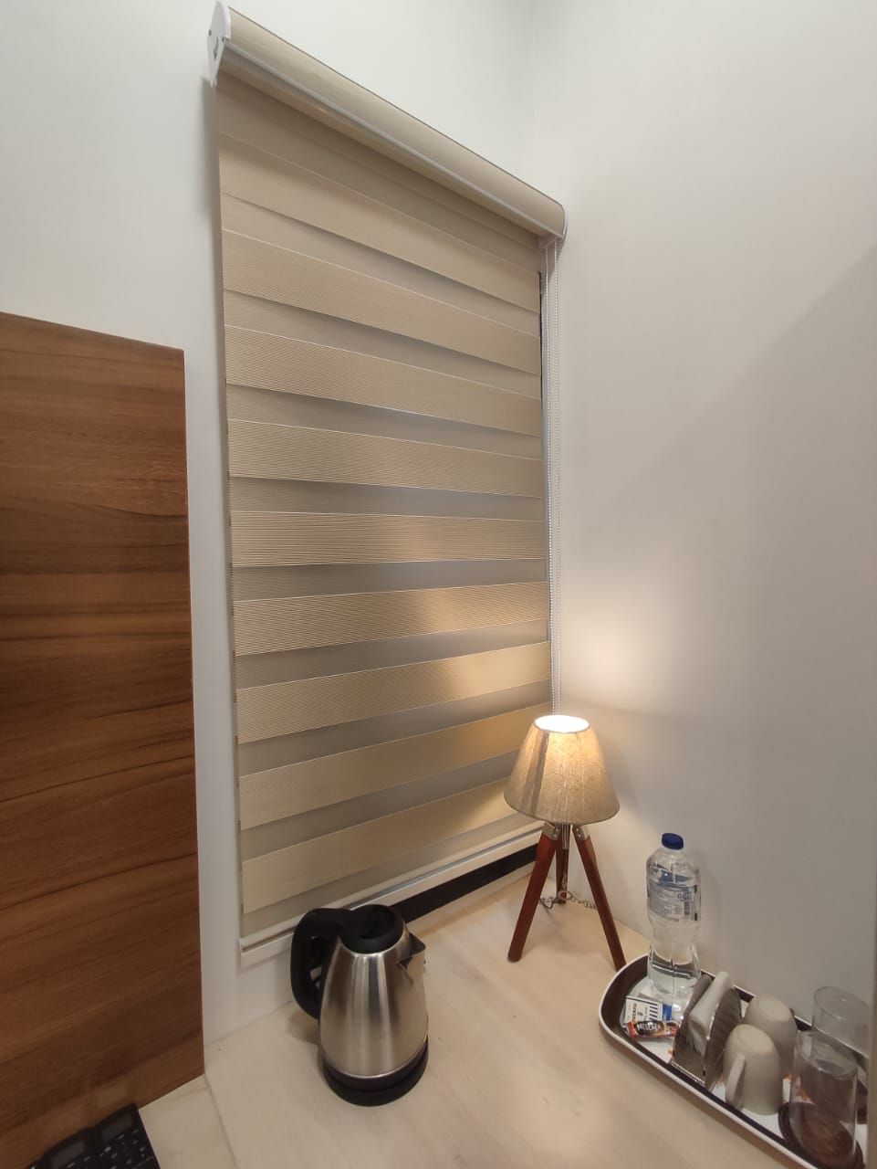 Une chambre d’hôtel avec un plancher en bois, une bouilloire, un plateau avec une bouteille d’eau, une lampe et une fenêtre avec des stores.