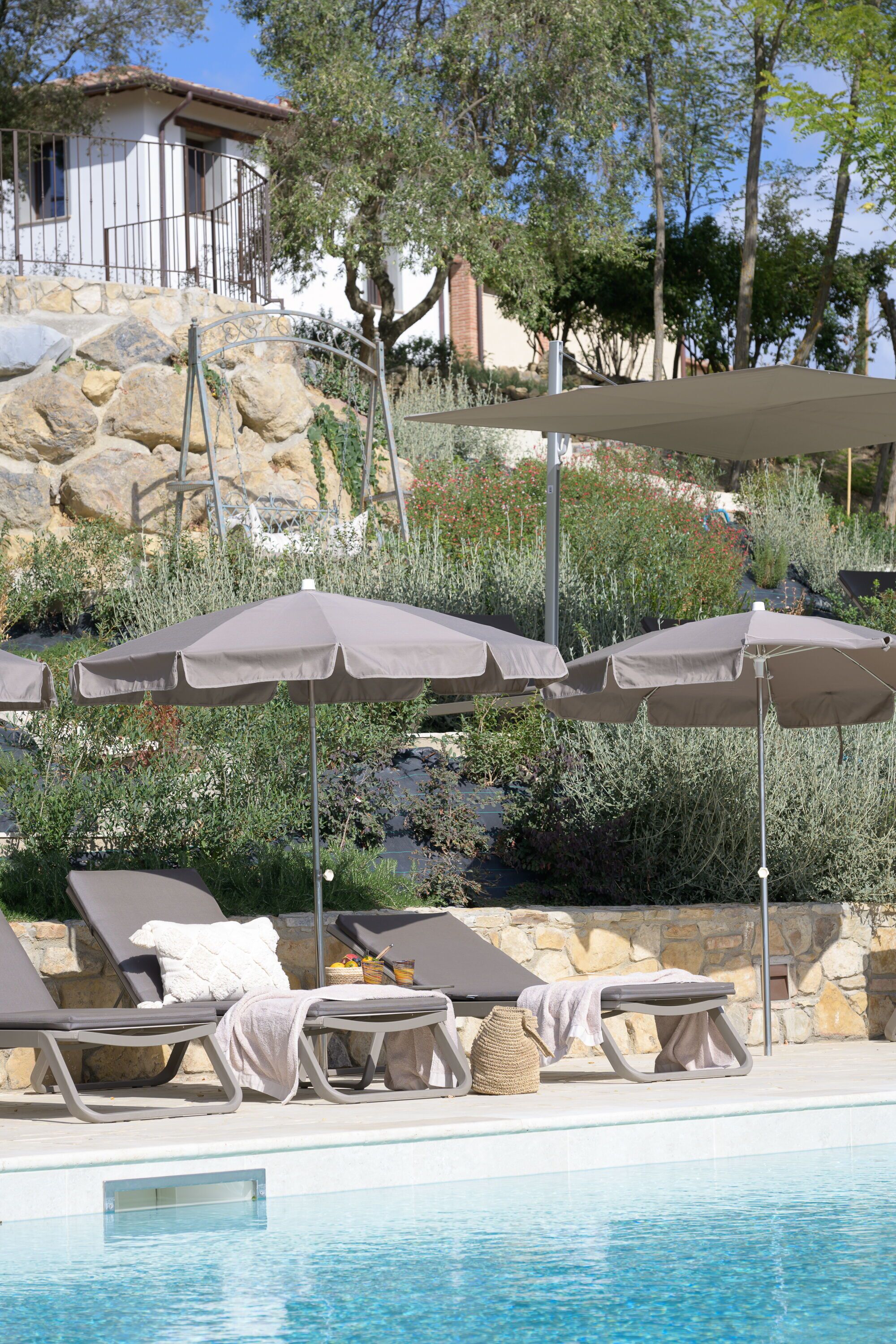 Un espace piscine agrémenté de chaises longues et de parasols, entouré d’un mur de pierre et de végétation.