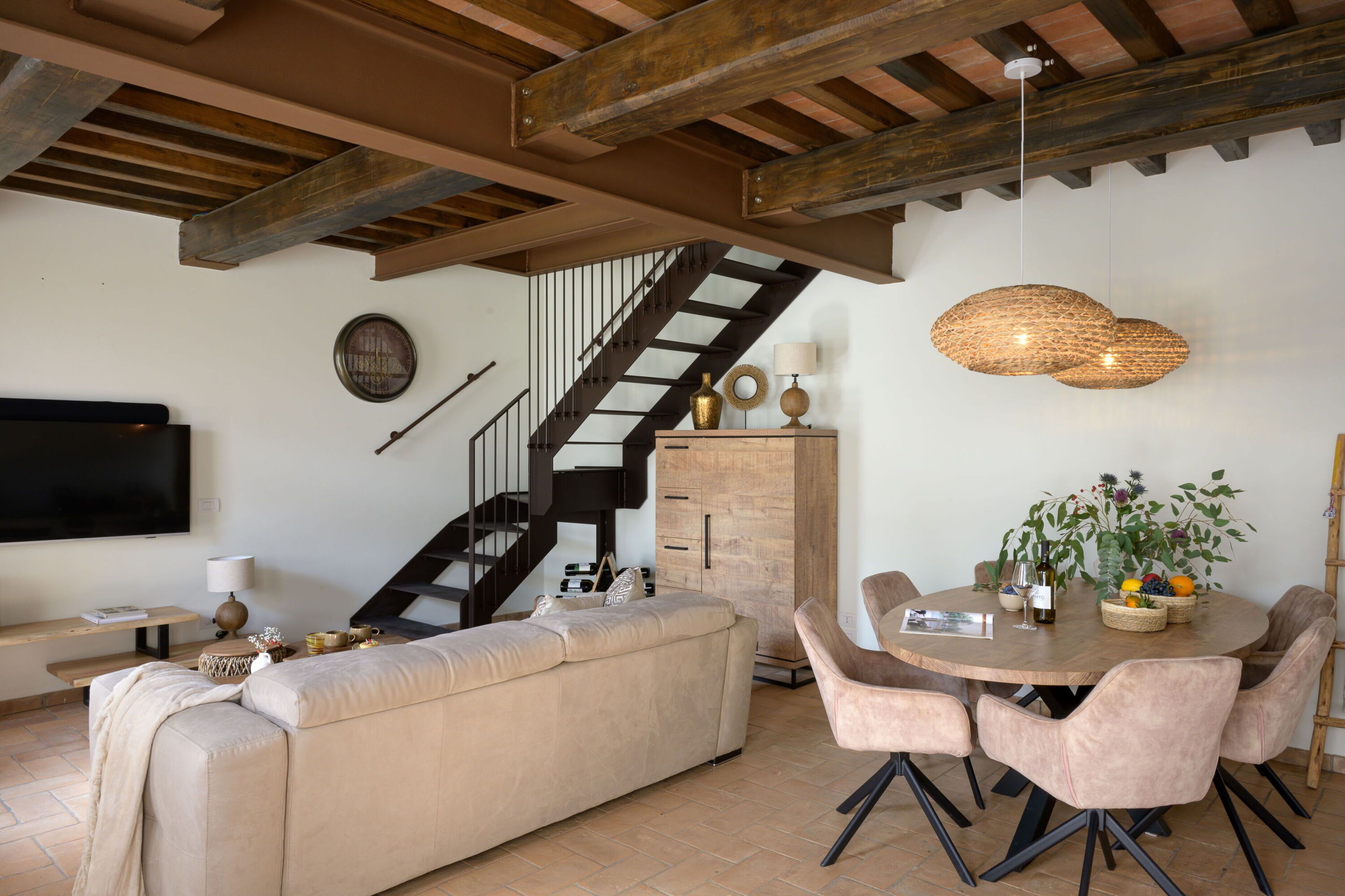 Een moderne woonkamer met een bank, eettafel, stoelen, een trap en een aan de muur bevestigde televisie.