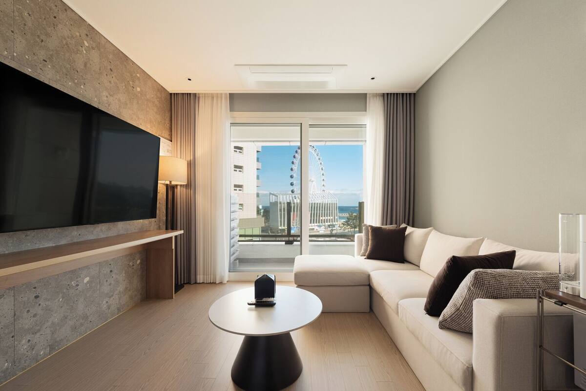 Ein modernes Hotelzimmer mit einem großen Flachbildfernseher an einer strukturierten Wand, einem runden Couchtisch, einem beigen Sofa mit Kissen und Blick auf ein Riesenrad durch eine Glastür.
