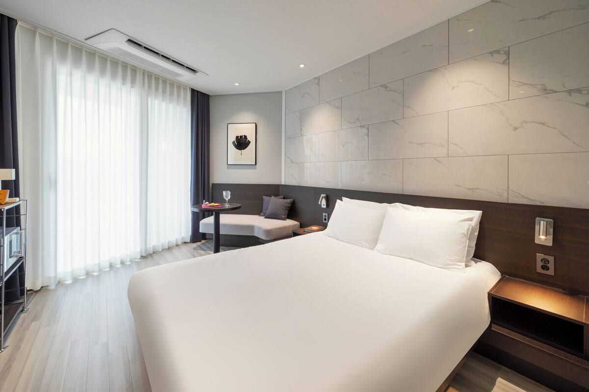 Ein modernes Hotelzimmer mit einem großen Bett, einem Nachttisch, einem kleinen Schreibtisch und einem Sessel.