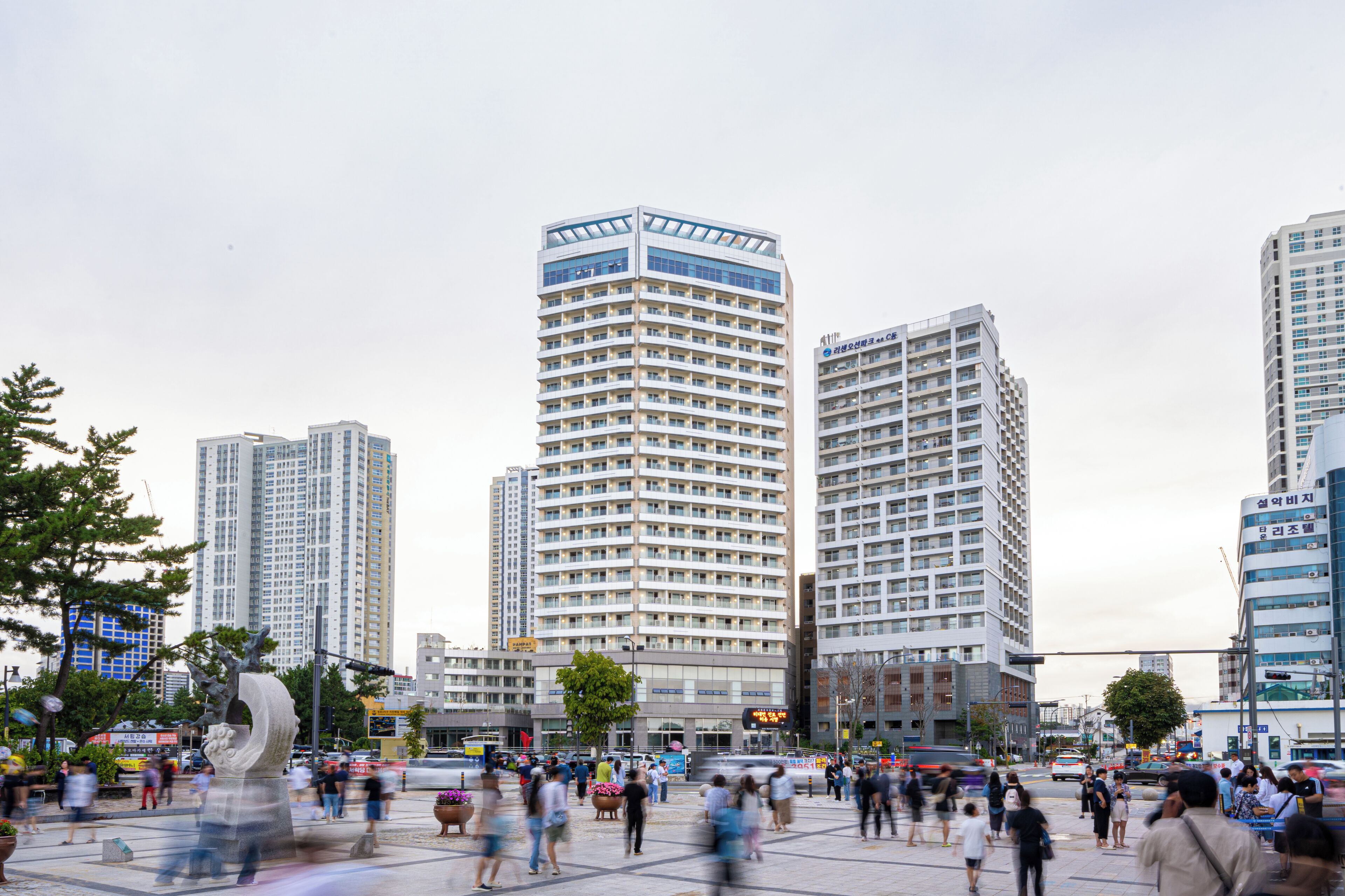 Una animada plaza urbana con modernos edificios altos, una escultura central y una multitud activa.