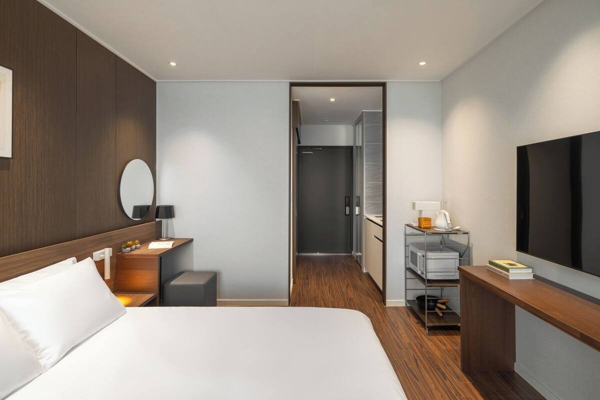 Ein modernes Hotelzimmer mit einem großen Bett, einem Nachttisch mit Lampe, einem kleinen Hocker, einem Schreibtisch mit Lampe, einem Fernseher und einem begehbaren Kleiderschrank.