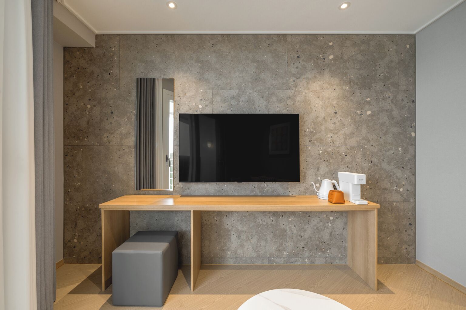 Una sala moderna con un televisor de pantalla plana grande montado en una pared texturizada, una mesa consola de madera con un hervidor y una cafetera, y un otoman gris.