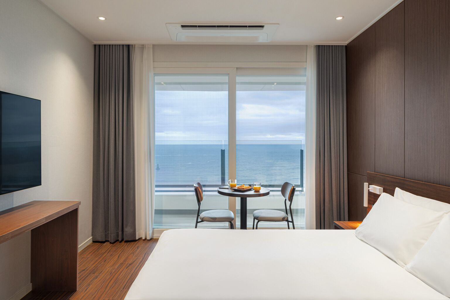 Ett hotellrum med en stor säng, en TV, en balkong med bord och stolar samt utsikt över havet.